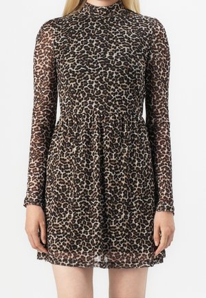 Femme portant une robe courte à manches longues et col haut, avec un motif léopard marron et noir, sur fond blanc.