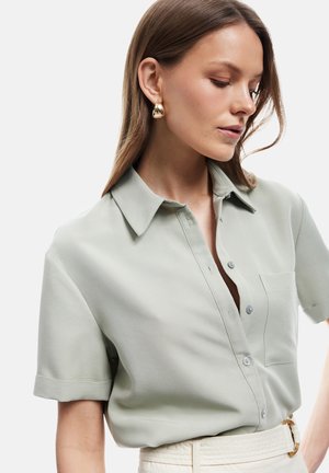 Kobieta o długich brązowych włosach, ubrana w jasnoszarą, krótką koszulę zapinaną na guziki i duże złote kolczyki w kształcie kół, patrząca w dół, z ręką na biodrze.