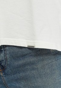 Camiseta blanca de algodón con dobladillo recto y una pequeña etiqueta metálica que dice "REPRESENT", combinada con jeans de mezclilla azul texturizados con costuras visibles.