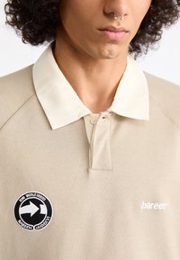 Joven con cabello rizado que lleva una camisa polo beige con cuello blanco, piercing en el septum y un parche en blanco y negro de "Serie Mundial 2016".