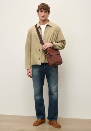 Fiatal férfi bézs dzsekiben, fehér ingben, kék farmerral, barna tornacipőben, és barna bőr keresztpántos táskát visel sötét pánttal.