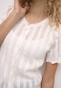 Vit stickad cardigan med ett ribbat mönster, korta puffärmar och rund halsringning, med knappstängning och texturerade detaljer.