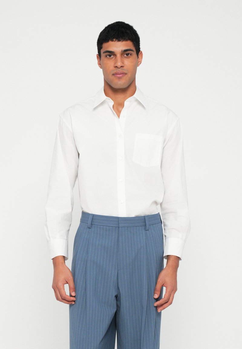 Weißes Langarmhemd mit Knopfleiste am Kragen und Brusttasche; kombiniert mit blauen, fein gestreiften Hosen mit geradem Schnitt.