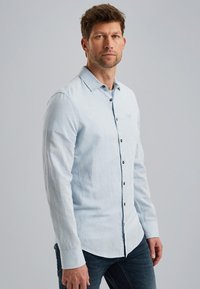 Camisa de manga larga de color azul claro, hecha de una tela texturada, con un cuello de botón y botones oscuros en la parte delantera. Usada con jeans oscuros.