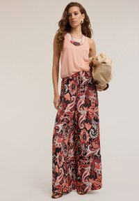 Spodnie w szerokim kroju z kwiatowym wzorem w kolorach czarnym, czerwonym i tealowym, zestawione z jasnoróżową, bezrękawnikową bluzką. Modelka trzyma beżową torbę z futrzanymi akcentami.