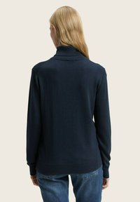 Pull en tricot bleu marine avec un col montant et des poignets côtelés, présentant une texture lisse et une coupe décontractée, porté sur un jean en denim.