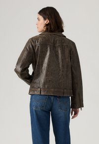 Brown leather jacket with a textured finish, cropped design, and prominent shoulder seams. Features a zippered cuff and back yoke detailing.