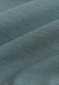 Textured, soft fabric in a muted blue-green color, displaying a woven pattern and subtle variations in shading. Smooth surface with gentle folds.