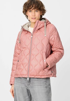 Frau trägt eine hellrosa gesteppte Kapuzenjacke mit Kordelzug, durchgehendem Reißverschluss vorne und graue Jeans vor weißem Hintergrund.