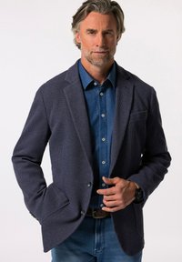 Granatowa marynarka o teksturowanej strukturze z klapami, z jedynym zapięciem na guzik i detalem w postaci trzech guzików przy mankietach, noszona na niebieskiej koszuli dżinsowej.