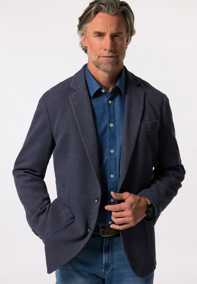 Granatowa marynarka o teksturowanej strukturze z klapami, z jedynym zapięciem na guzik i detalem w postaci trzech guzików przy mankietach, noszona na niebieskiej koszuli dżinsowej.