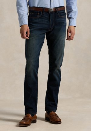 Homme portant un jean bleu foncé, une ceinture en cuir marron, une chemise bleue claire boutonnée et des mocassins en cuir marron, debout sur un sol beige.