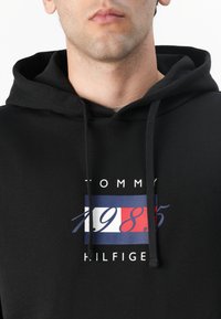 Sort hættetrøje lavet af bomuld, med et frontgrafik i blå, rød og hvid med teksten "TOMMY 1985 HILFIGER" og snorehætte.