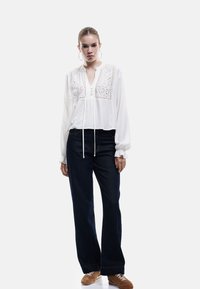 Hvid bluse med broderede detaljer, knaplukning og snoretræk ved halsen, parret med mørkeblå brede jeans og brune sneakers.