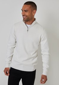 Weißer Pullover mit Viertel-Reißverschluss-Kragen, gerippten Bündchen und Saum, aus einem weichen Strickstoff. Getragen über einem weißen Hemd und schwarzen Hosen.