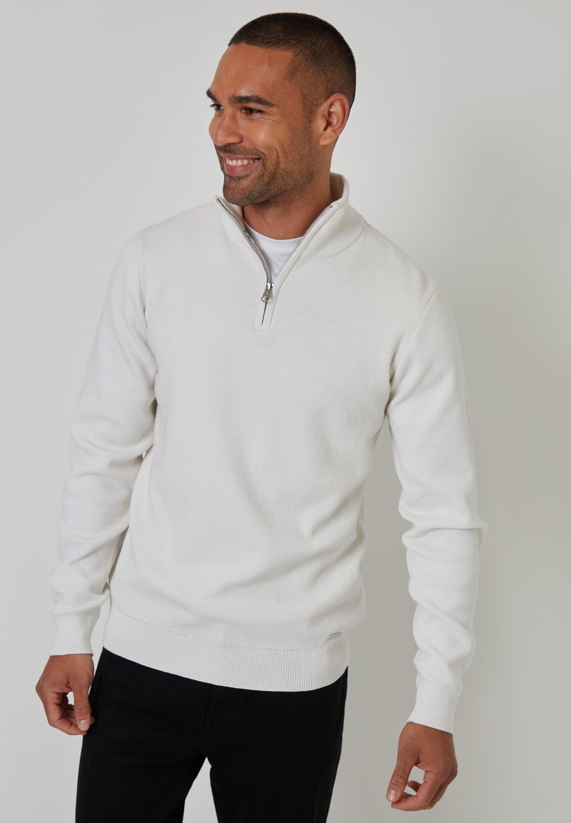 Weißer Pullover mit Viertel-Reißverschluss-Kragen, gerippten Bündchen und Saum, aus einem weichen Strickstoff. Getragen über einem weißen Hemd und schwarzen Hosen.