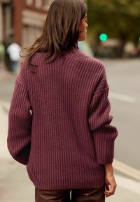 Übergroßer, gerippter Strickpullover in tiefem Burgunderrot mit einem hohen Kragen und fallenden Schultern, der über braunen Lederhosen liegt.