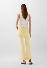 Jeans evazate de culoare galben deschis, cu margine ciufulită, două buzunare spate cu cusături în formă de diamant, asortate cu un maieu alb cu textură ribbed.