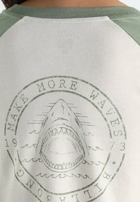 T-shirt bianco in cotone con maniche raglan verdi. Presenta una grafica di squalo con il testo "Fai più onde" circondato sotto.