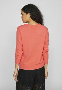 Korallfarget strikkegenser med rund hals og ribbestrikkede mansjetter, kombinert med svarte spisse bukser med en teksturert mønster.