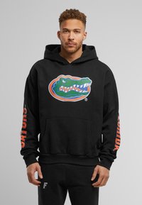 Sudadera negra de felpa de algodón con un gran logo de cocodrilo verde en la parte frontal. Texto naranja que recorre la manga izquierda. Bolsillo tipo canguro.