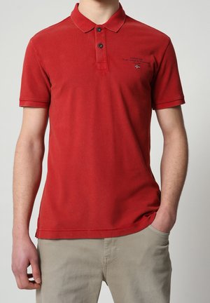 Rød polo skjorte med korte ærmer, knapper og lille logo på brystet, båret af en person med den ene hånd i lommen på beige bukser.