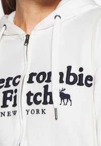Bílá zipová mikina vyrobená z měkké tkaniny, která má vyšívaný námořnický modrý text "Abercrombie Fitch" a logo losa, s akcenty na šňůrkách.