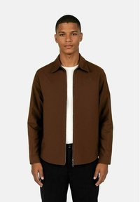 Brun zip-front jakke med spiss krage, lange ermer, og teksturert stoff over en hvit skjorte, paret med svarte bukser.