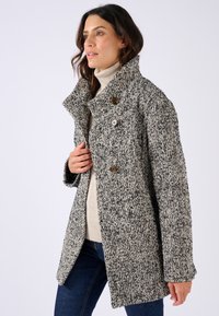 Manteau grisonnant et noir à motifs, avec un large col, des boutons à l'avant et une coupe ample. Porté sur un léger pull et associé à un jean foncé.