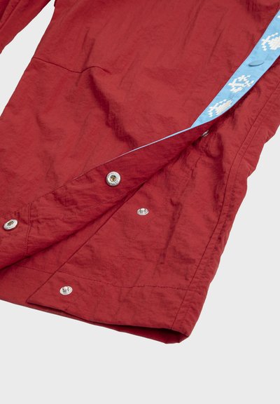 Vêtement rouge avec boutons-pression argentés partiellement ouverts, révélant une bordure bleu clair ornée de motifs géométriques blancs.