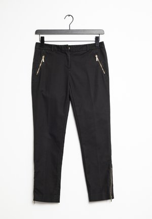 Zwarte broek van geweven stof, met gouden ritszakken, slim fit en zijritsen bij de enkel. Hangt aan een zwarte haak.