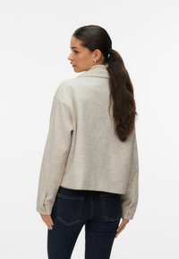 Femme avec une longue queue de cheval noire, portant une veste beige clair et un jean bleu foncé, debout et regardant vers sa gauche contre un fond uni.