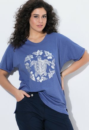 Kobieta z kręconymi włosami, ubrana w niebieski T-shirt z motywem srebrnej żółwi morskiej i kwiatowym, w połączeniu z ciemnymi spodniami, pozująca z jedną ręką w kieszeni.