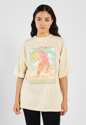 Beige oversize tričko s grafikou tygra, slunce a měsíce se textem "STAY WILD" v modrozelené barvě. Vyrobeno z bavlny s hladkou texturou.