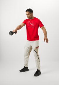 Rød t-shirt med korte ærmer og logo, lyse beige bukser med sidelommer og lynlåse, samt sorte sneakers, med en person der løfter en håndvægt.