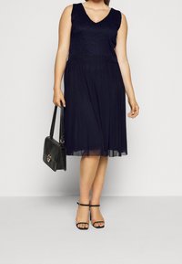 Marinblå klänning med spetsliv, V-ringning och flödig chiffonskjol. Modellen håller en svart texturerad handväska och bär svarta sandaletter.