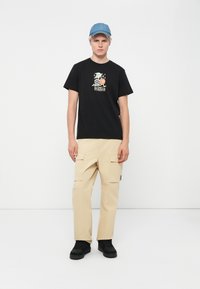Black cotton t-shirt with graphic print and text, beige cargo pants with side pockets, and black sneakers. Denim cap completes the look.