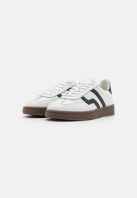 White leather sneakers featuring black side stripes, round toe, flat brown rubber sole, and designed with white laces and stitching details.