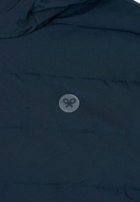 Giacca imbottita di colore blu scuro, con design a righe orizzontali e una toppa circolare grigia con un'illustrazione di forbici.