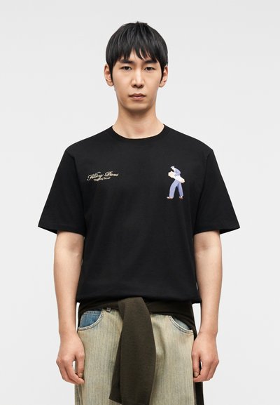 Μαύρο βαμβακερό t-shirt με εκτύπωση μπροστά που απεικονίζει έναν χαρακτήρα με μπλε ντύσιμο και το κείμενο «Killing Bees Playing Bear». Κοντά μανίκια, άνετη εφαρμογή.