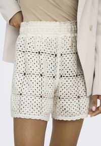 Shorts all'uncinetto color crema con un motivo testurizzato, orlo ondulato e vita elastica, indossati con un top aderente e un blazer leggero.