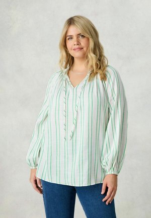 Blouse blanche à rayures verticales vertes, avec un col en V, un cordon de serrage au niveau du cou et des manches bouffantes élastiques. Associée à un jean bleu.