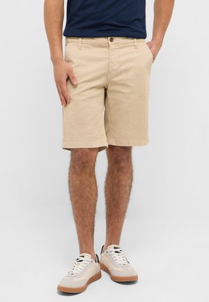 Mann in beige knielangen Shorts, navyblauem Hemd und weißen Sneakers, stehend mit den Händen in den Taschen vor weißem Hintergrund.