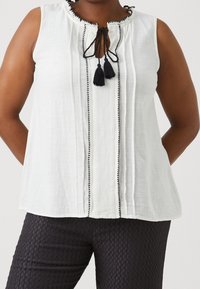 Bílý bezrukávový top s volánkovým výstřihem, černými třásněmi a kontrastním černým lemem vpředu; nošený s texturovanými černými kalhotami.