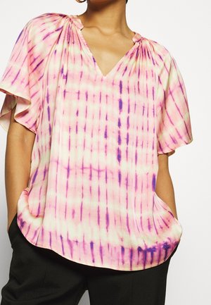 Mujer con blusa de manga corta, suelta, de teñido anudado rosa y púrpura con escote en V fruncido y pantalones negros, con las manos en los bolsillos.