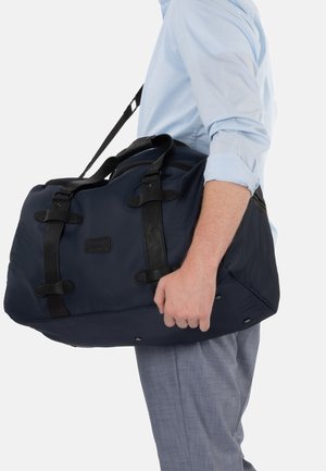 Man wearing light blue shirt and gray pants holding navy blue duffel bag with black straps by the handles at his side.