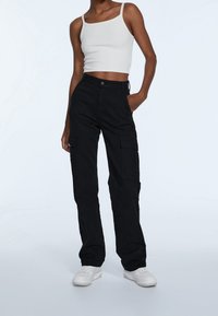 Schwarze Cargo-Hosen mit mehreren Taschen, hoher Taille und geradem Schnitt. Getragen mit einem weißen, verkürzten Tank-Top und weißen Sneakers.