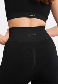 Schwarzes Sport-BH- und Leggings-Set mit hohem Bund. Der Stoff ist glatt und hat eine enganliegende Passform. Das "OCEANSAPART"-Logo ist auf beiden Teilen sichtbar.