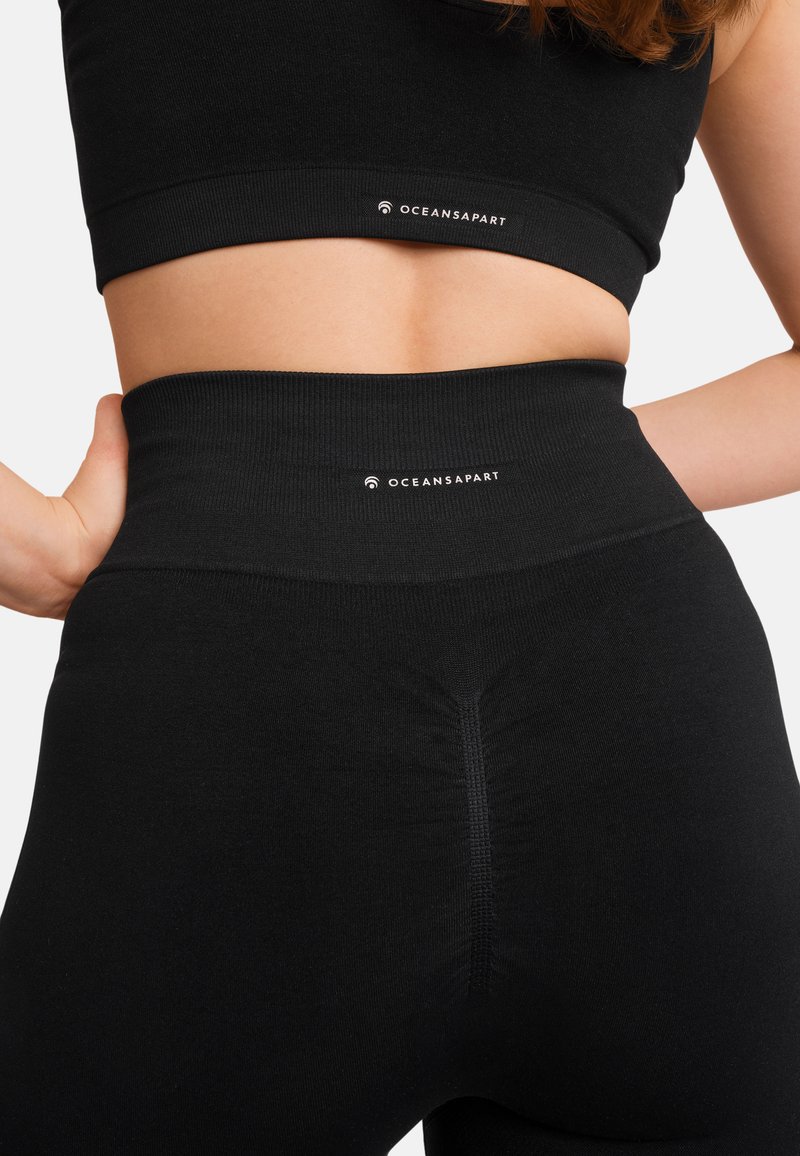 Czarny zestaw sportowy składający się z biustonosza i legginsów, z wysokim stanem. Materiał jest gładki, a krój dopasowany. Logo "OCEANSAPART" widoczne na obu elementach.
