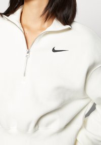 White sweatshirt with a quarter zip, ribbed cuffs, and hem. Features a small black Nike logo on the left chest and a soft, textured fabric.